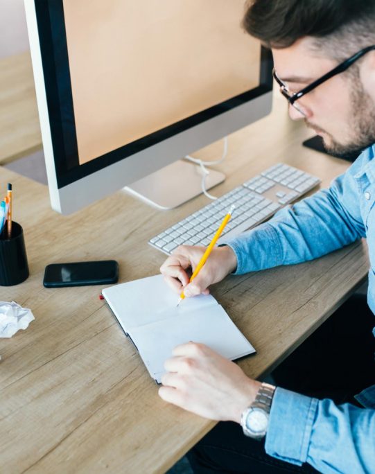 young-attractive-man-glassess-is-working-his-workplace-office-he-wears-blue-shirt-he-is-writing-notebook-view-from